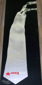 Indulge in the allure of fifty shades of grey with this custom aisle runner design. A silk grey tie, painted in a large-scale fashion on a caviar black runner, exudes sophistication and sensuality. This sleek and sultry design provides the perfect canvas for the addition of the "Sexy Hair" logo, making it an ideal choice for a special event.